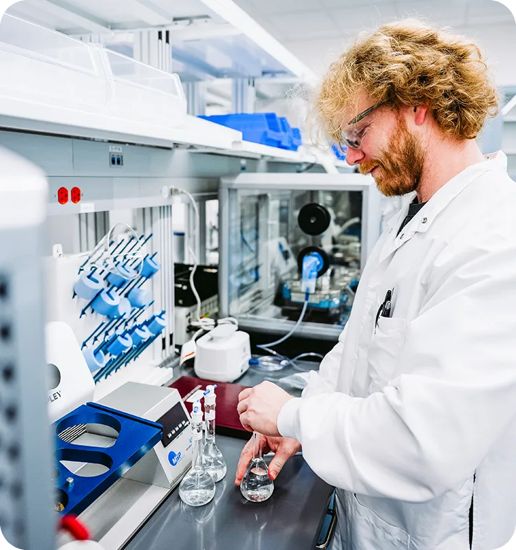 A scientist in a white lab coat and safety glasses prepares samples in volumetric flasks at a lab bench. Analytical instruments and supplies fill the lab around him.