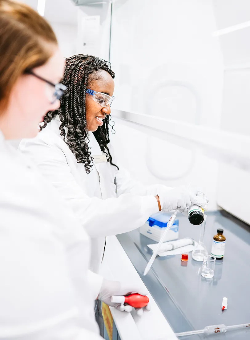 Two scientists in white lab coats, safety glasses and gloves work at a lab bench. One of them measures liquid with a pipette next to an amber reagent bottle, small beakers and a flask.