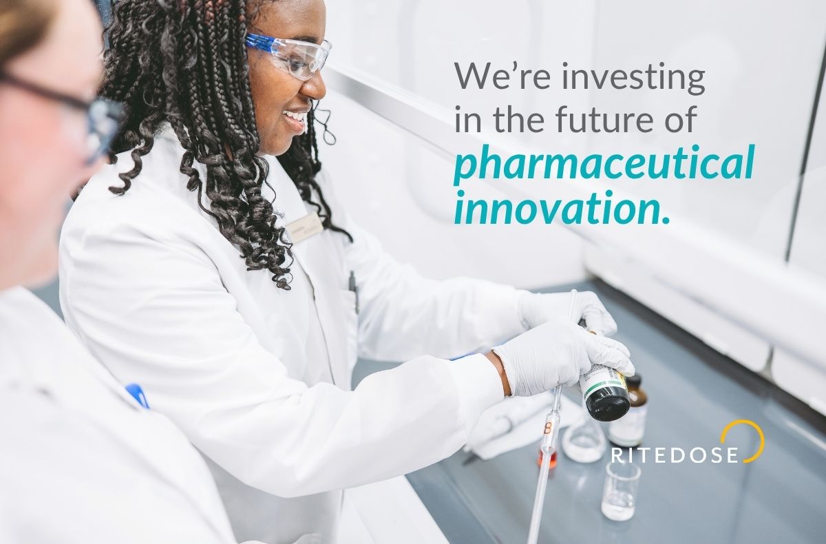 A smiling scientist in a lab coat, safety glasses, and gloves pours from a reagent bottle into a burette at a laboratory fume hood while a colleague looks on. Overlay text reads "We're investing in the future of pharmaceutical innovation" with the Ritedose logo.