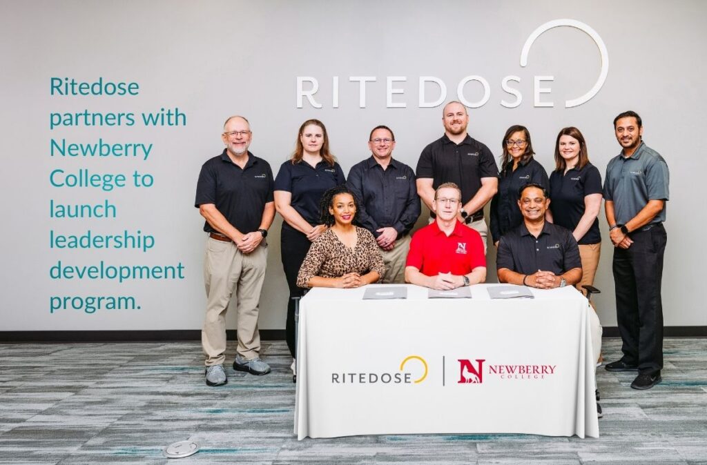 Ritedose and Newberry College representatives pose for a group photo, with three seated at a Ritedose-branded signing table and seven standing behind them in front of a large Ritedose logo wall. Overlay text reads “Ritedose partners with Newberry College to launch leadership development program.”